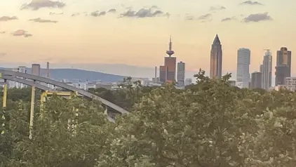 Skyline von Frankfurt am Main bei Sonnenuntergang, mit markanten Hochhäusern und Fernsehturm im Hintergrund. Im Vordergrund sind Bäume und ein Teil einer Achterbahn zu sehen.