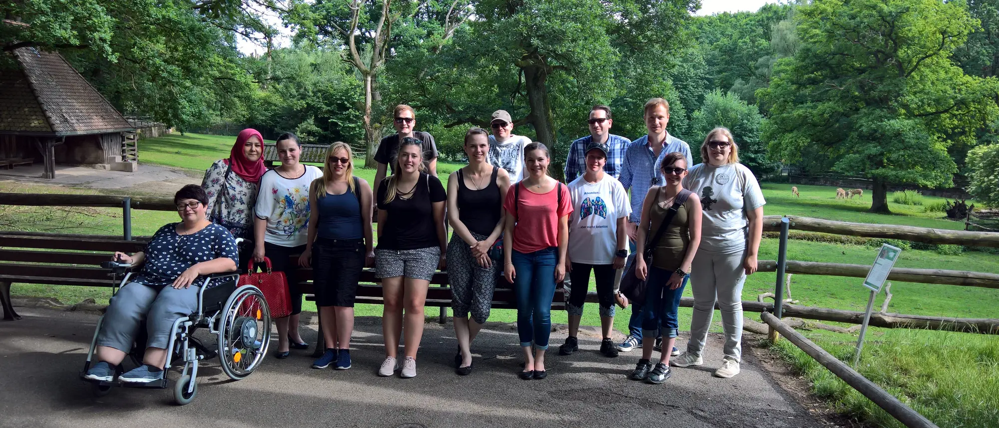 Auf dem Bild sind Menschen in einer freundlichen Gruppe versammelt. Einige tragen bunte T-Shirts, während sie in einer natürlichen Umgebung stehen. Der Hintergrund zeigt einen sonnigen Park mit einer Wiese und Tieren.