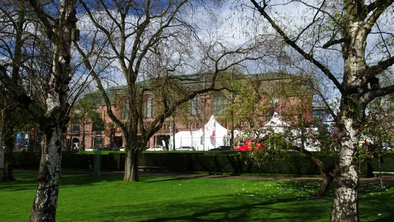 Das Bild zeigt das Kongresszentrum Rosengarten. Im Vordergrund sind Bäume und eine grüne Rasenfläche zu sehen. Im Hintergrund steht das Gebäude des Kongresszentrums aus rötlichem Stein. Vor dem Gebäude sind weiße Zelte und einige Fahrzeuge sichtbar.