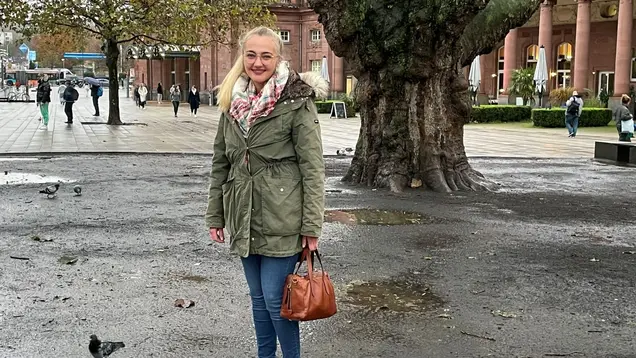 Das Bild zeigt Lara während eines Treffens in Wiesbaden. Sie steht auf einem Platz, umgeben von herbstlichen Bäumen. Lara trägt eine grüne Jacke und einen Schal und hält eine braune Handtasche in der Hand. Im Hintergrund sind Passanten und Gebäude zu sehen, die eine angenehme urbane Atmosphäre schaffen.