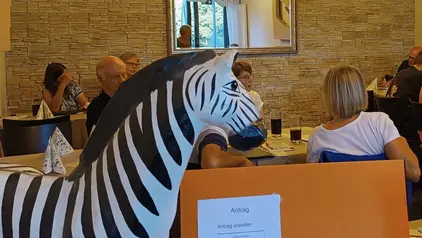 Ein Pappmodell eines Zebras steht im Vordergrund vor einer Gruppe von Menschen, die an einem Tisch in einem Raum mit Steinwand sitzen.