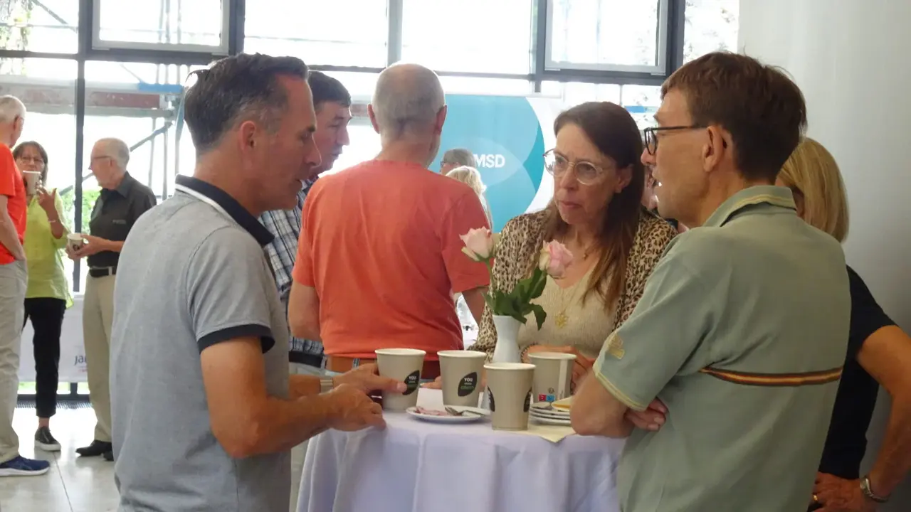 Eine Gruppe von Menschen steht an einem Stehtisch und unterhält sich während einer Veranstaltung. Auf dem Tisch stehen Kaffeebecher und eine Vase mit rosa Rosen. Im Hintergrund sind weitere Personen zu sehen, die sich ebenfalls unterhalten. Der Raum ist hell und modern gestaltet, mit großen Fenstern, durch die Tageslicht hereinfällt.