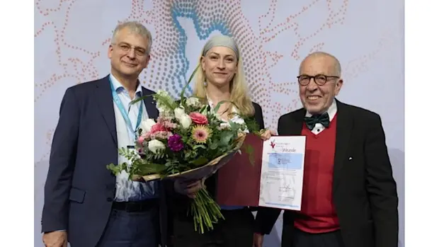 Drei Personen bei der Verleihung eines Forschungspreises: Die Preisträgerin in der Mitte hält einen Blumenstrauß und eine Urkunde, flankiert von zwei männlichen Kollegen, alle lächeln in die Kamera.