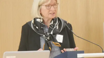 Eine Frau mit blonden Haaren und Brille spricht an einem Podium. Sie trägt einen schwarzen Blazer und einen gemusterten Schal, vor ihr ist ein Mikrofon positioniert.