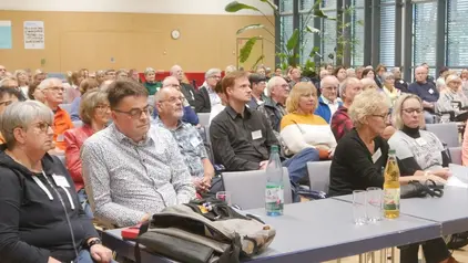 Eine Gruppe von Menschen sitzt aufmerksam in einem Konferenzraum. Im Vordergrund stehen Getränke auf den Tischen, im Hintergrund sind Poster und Pflanzen zu sehen.