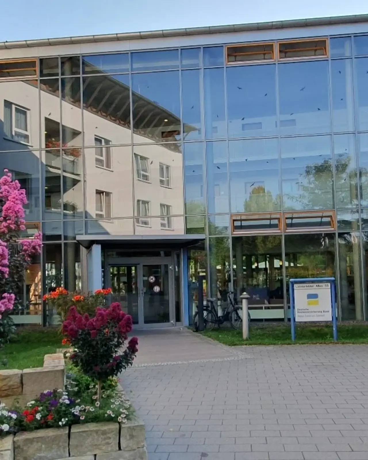 Das Bild zeigt den Eingang eines modernen Gebäudes mit großen Glasfenstern. Vor dem Eingang blühen pinke und lila Blumen, die dem Bild Farbe und Leben verleihen. Im Hintergrund spiegelt sich ein weiteres Gebäude in den Glasfenstern, und der Himmel ist klar und sonnig.