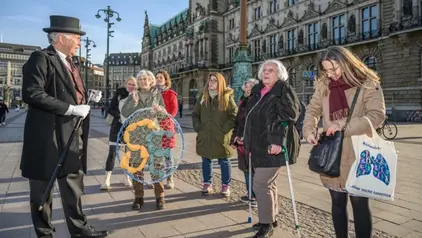 Eine Gruppe von Menschen steht auf einem Platz vor einem historischen Gebäude. Ein Mann in einem schwarzen Frack und Zylinderhut hält einen Spazierstock und spricht zur Gruppe. Eine Frau hält ein Netz mit bunten Stoffteilen, während eine andere Frau eine Tasche trägt. Einige Personen tragen Winterjacken und hören aufmerksam zu.