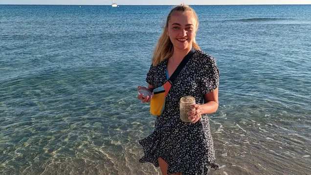 Das Bild zeigt Lara an einem Strand auf Mallorca. Sie steht barfuß im flachen Wasser, trägt ein Sommerkleid und hält ein Glas mit Sand in der Hand. Im Hintergrund ist das Meer mit einem Segelboot zu sehen. Der klare Himmel und das ruhige Wasser schaffen eine entspannte, sommerliche Atmosphäre.