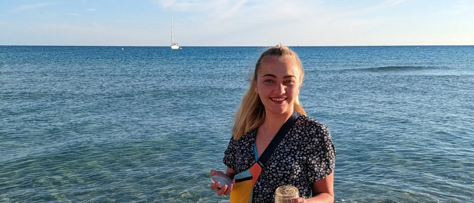 Das Bild zeigt Lara an einem Strand auf Mallorca. Sie steht barfuß im flachen Wasser, trägt ein Sommerkleid und hält ein Glas mit Sand in der Hand. Im Hintergrund ist das Meer mit einem Segelboot zu sehen. Der klare Himmel und das ruhige Wasser schaffen eine entspannte, sommerliche Atmosphäre.