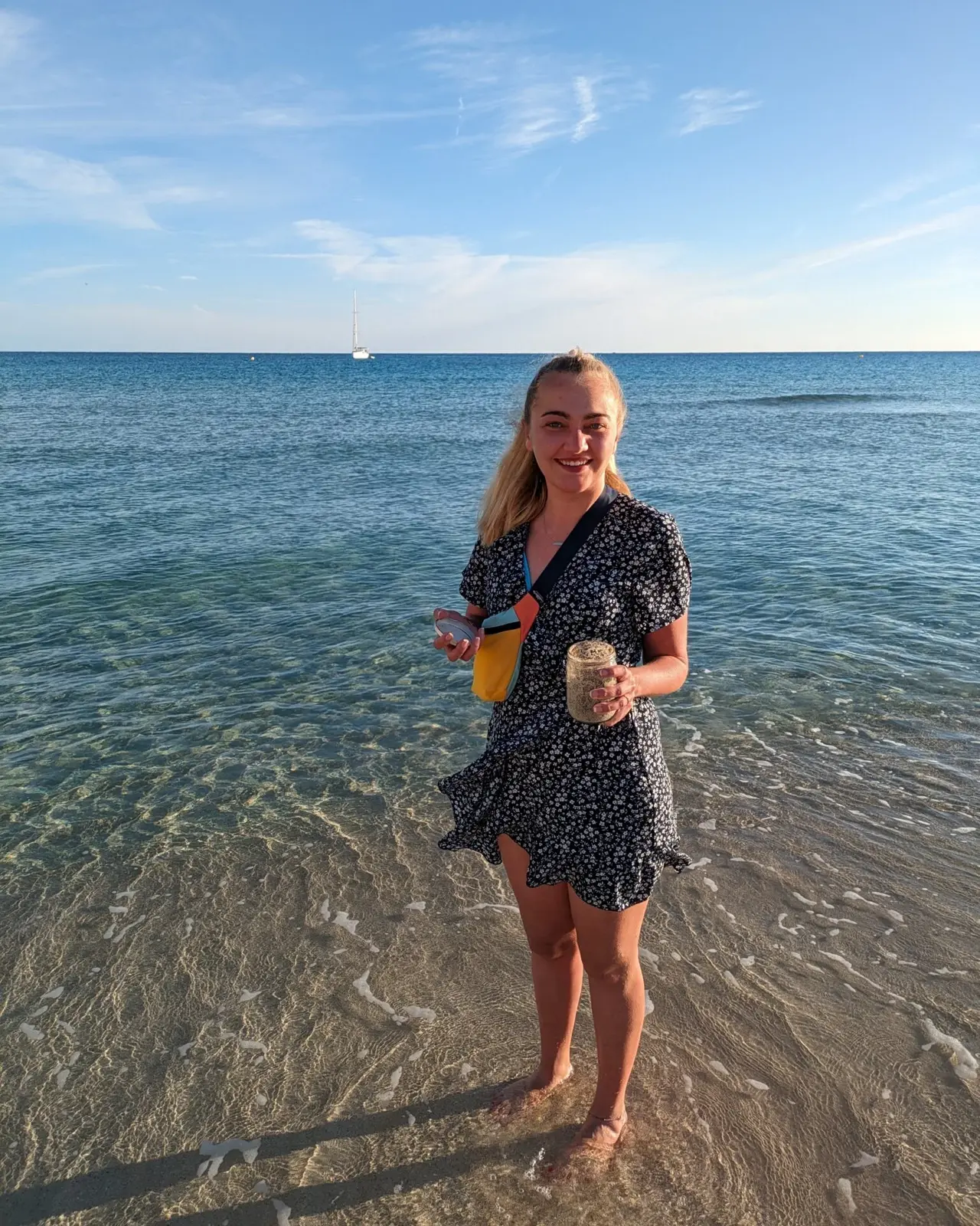 Das Bild zeigt Lara an einem Strand auf Mallorca. Sie steht barfuß im flachen Wasser, trägt ein Sommerkleid und hält ein Glas mit Sand in der Hand. Im Hintergrund ist das Meer mit einem Segelboot zu sehen. Der klare Himmel und das ruhige Wasser schaffen eine entspannte, sommerliche Atmosphäre.