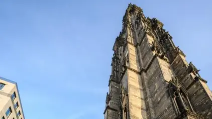 Ansicht einer imposanten, gotischen Kirchenruine aus einer niedrigen Perspektive, die den hohen Turm und die kunstvollen Steinmetzarbeiten hervorhebt. Die Kirche steht neben modernen Gebäuden, die im Hintergrund zu sehen sind. Der Himmel ist klar und blau, und im Vordergrund befindet sich eine Rasenfläche.