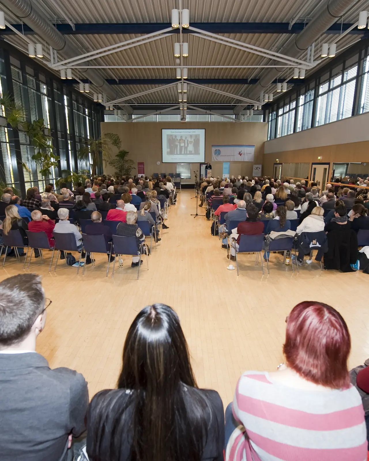 Das Bild zeigt einen großen, hellen Konferenzraum mit einer hohen Decke und einer Glasfront auf der linken Seite. Der Raum ist gefüllt mit zahlreichen Teilnehmern, die in mehreren Reihen auf Stühlen sitzen und einer Präsentation auf einer Leinwand an der Stirnseite des Raumes folgen.