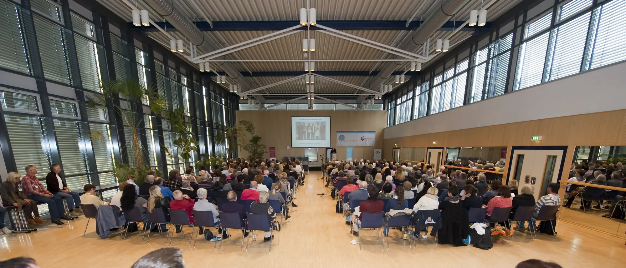 Das Bild zeigt einen großen, hellen Konferenzraum mit einer hohen Decke und einer Glasfront auf der linken Seite. Der Raum ist gefüllt mit zahlreichen Teilnehmern, die in mehreren Reihen auf Stühlen sitzen und einer Präsentation auf einer Leinwand an der Stirnseite des Raumes folgen.