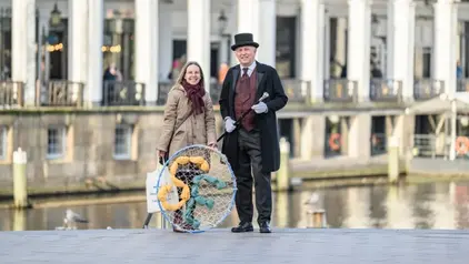 Eine Frau und ein Mann stehen draußen vor einem historischen Gebäude mit weißen Bögen. Die Frau trägt einen beigen Mantel und einen Schal und hält ein Netz mit bunten Stoffteilen. Der Mann trägt einen schwarzen Frack, einen Zylinderhut und hält einen Spazierstock.