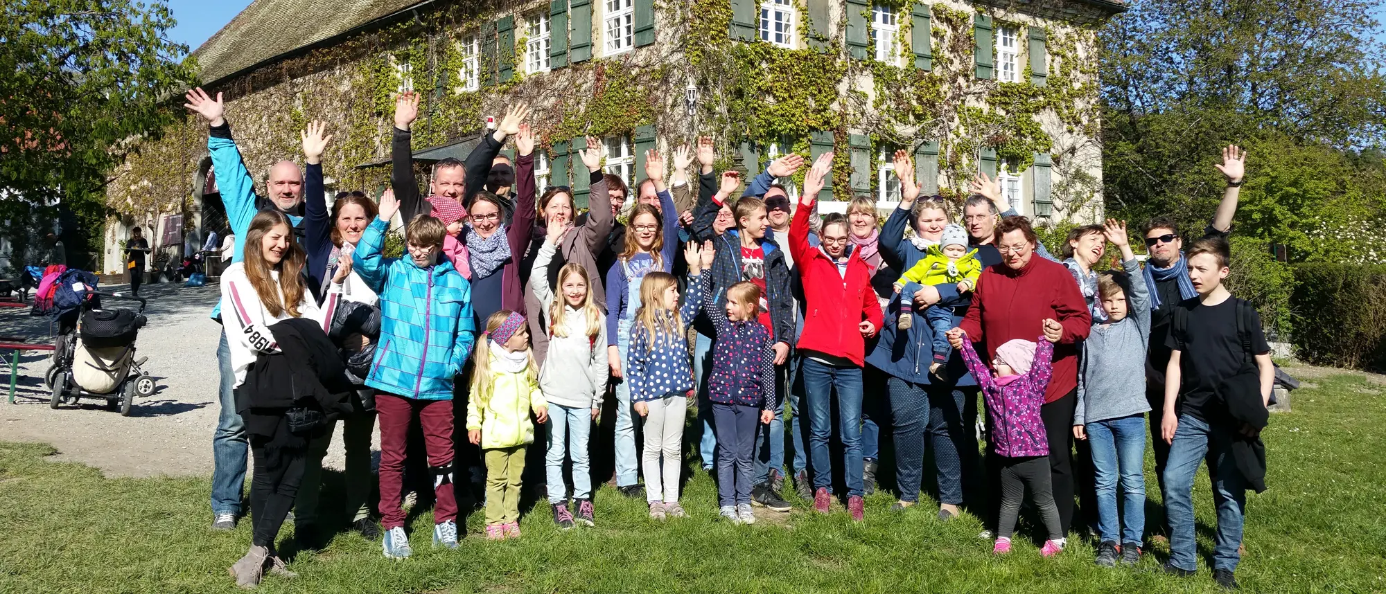 Gruppenfoto mit vielen Erwachsenen und Kindern, die vor einem mit Efeu bewachsenen historischen Gebäude stehen und in die Kamera winken. Die Stimmung ist fröhlich und das Wetter sonnig.