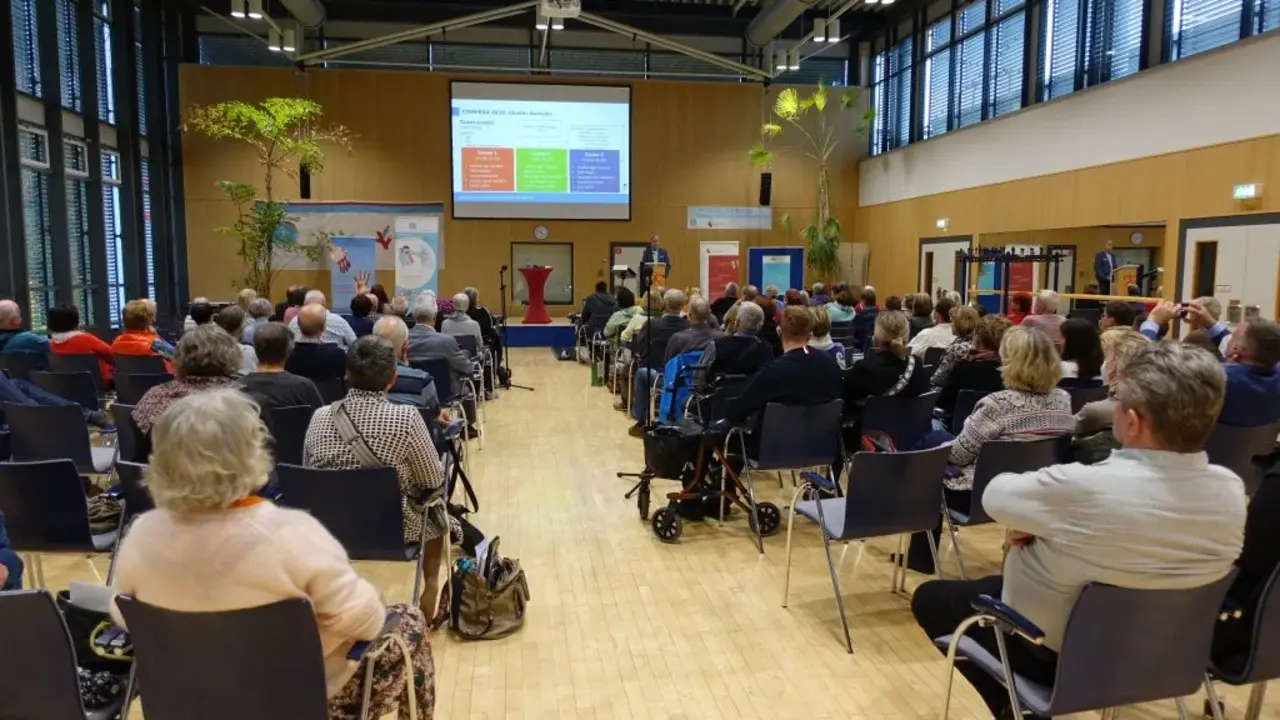 Ein Auditorium voll mit Menschen hört einer Präsentation zu, die auf einer großen Leinwand vorne im Raum gezeigt wird. Der Saal ist geräumig und mit Pflanzen dekoriert, während der Vortrag in einer professionellen Umgebung stattfindet.