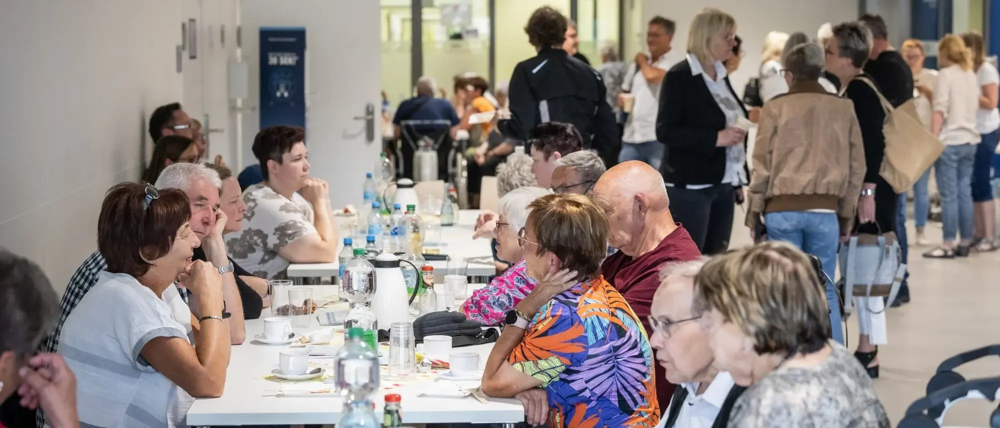 Eine Gruppe von Menschen sitzt an einem langen Tisch und unterhält sich. Im Hintergrund sind weitere Personen zu sehen, die in kleinen Gruppen stehen und sich ebenfalls unterhalten.