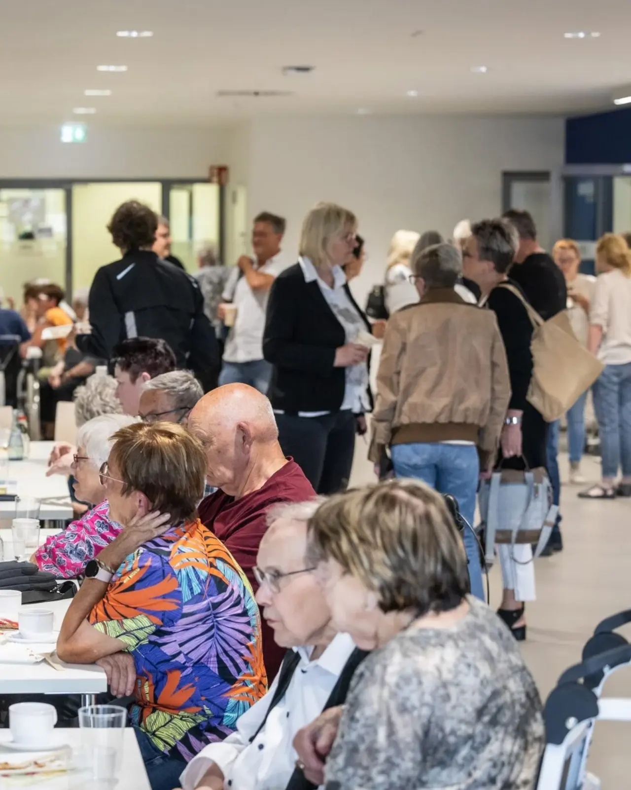 Eine Gruppe von Menschen sitzt an einem langen Tisch und unterhält sich. Im Hintergrund sind weitere Personen zu sehen, die in kleinen Gruppen stehen und sich ebenfalls unterhalten.