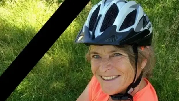 Helga Kuhn, eine lächelnde Frau, die einen Fahrradhelm und ein oranges T-Shirt trägt, sitzt im Gras. Sie hat kurze blonde Haare und eine freundliche Ausstrahlung. Ein schwarzer Trauerflor befindet sich in der linken oberen Ecke des Bildes.