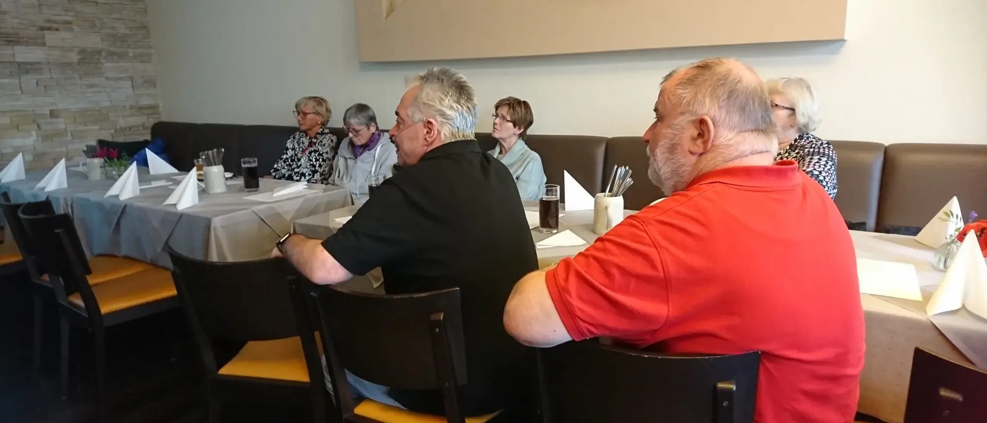 Mehrere Personen sitzen an einem gedeckten Tisch in einem Restaurant oder Veranstaltungsraum und hören aufmerksam zu. Der Tisch ist mit grauen Tischdecken, Gläsern und gefalteten Servietten dekoriert. Die Atmosphäre wirkt ruhig und konzentriert, während die Personen sich auf etwas außerhalb des Bildes fokussieren.