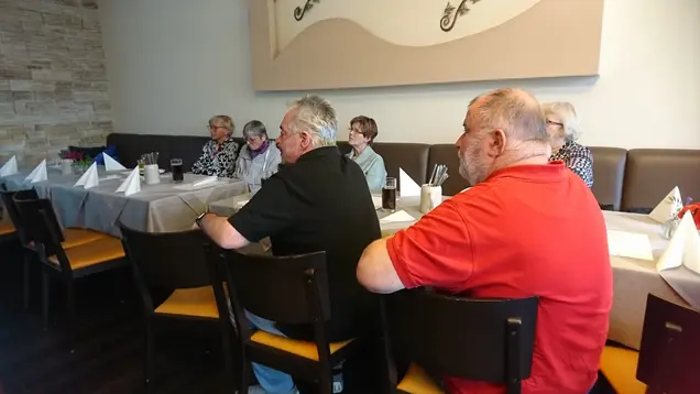 Mehrere Personen sitzen an einem gedeckten Tisch in einem Restaurant oder Veranstaltungsraum und hören aufmerksam zu. Der Tisch ist mit grauen Tischdecken, Gläsern und gefalteten Servietten dekoriert. Die Atmosphäre wirkt ruhig und konzentriert, während die Personen sich auf etwas außerhalb des Bildes fokussieren.