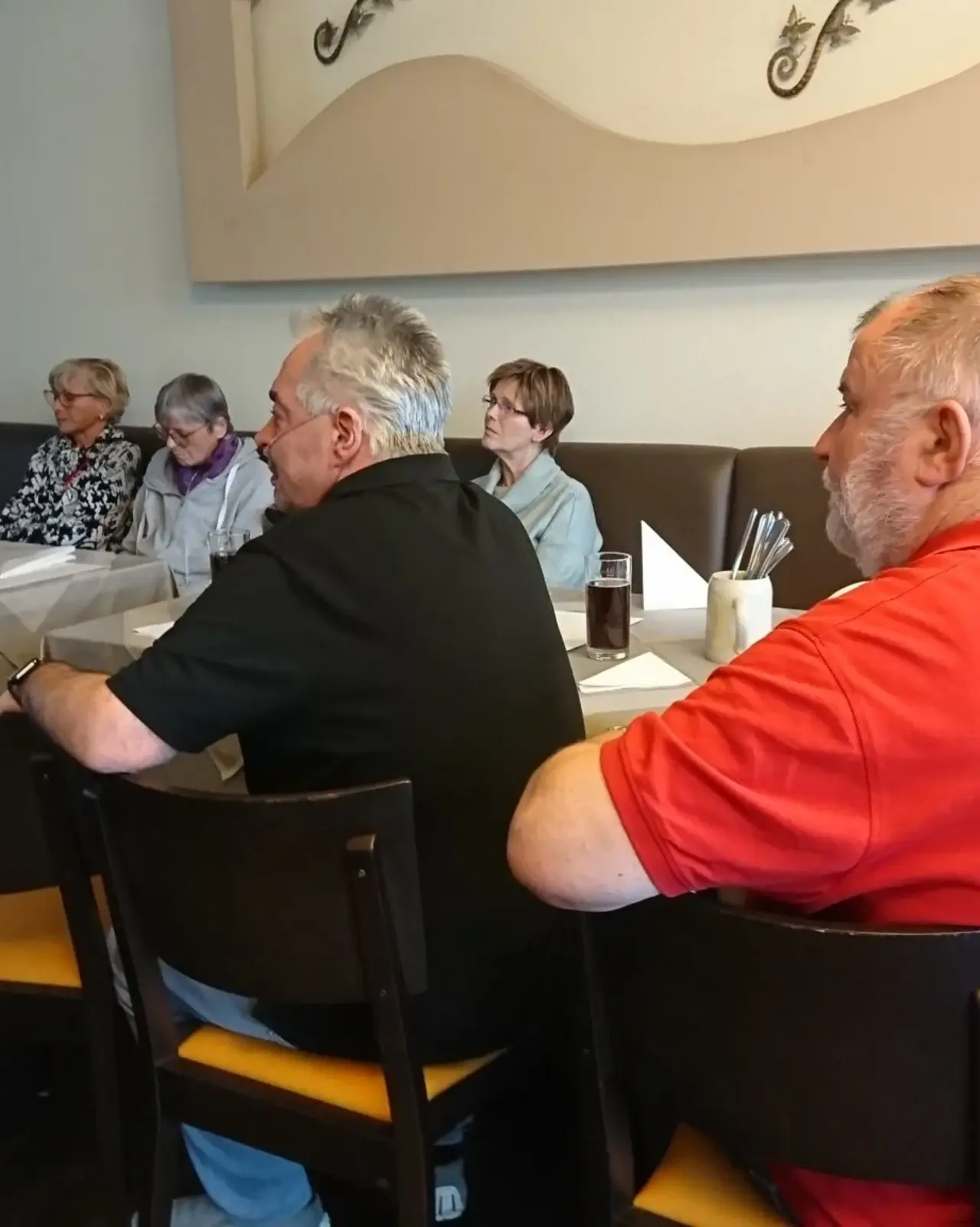 Mehrere Personen sitzen an einem gedeckten Tisch in einem Restaurant oder Veranstaltungsraum und hören aufmerksam zu. Der Tisch ist mit grauen Tischdecken, Gläsern und gefalteten Servietten dekoriert. Die Atmosphäre wirkt ruhig und konzentriert, während die Personen sich auf etwas außerhalb des Bildes fokussieren.