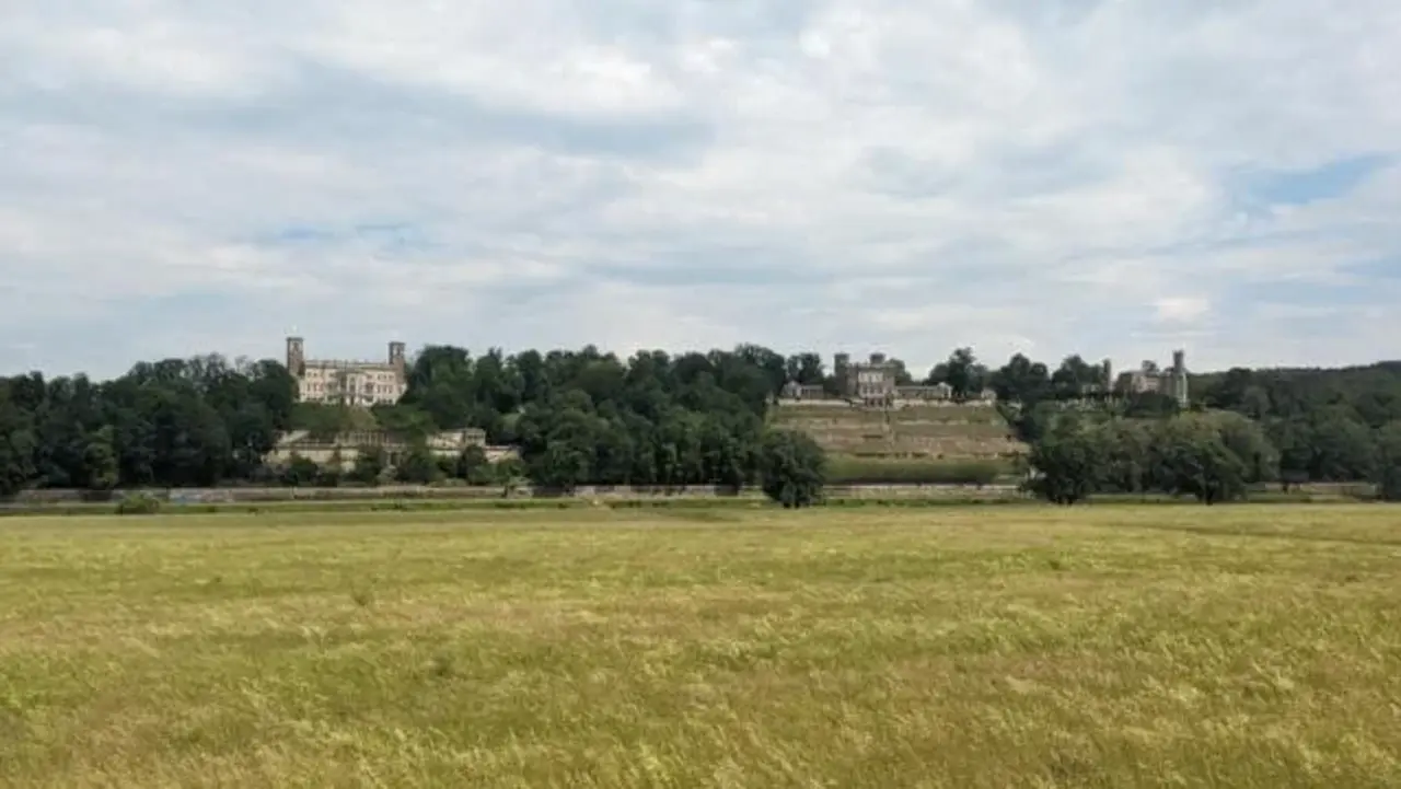 Weite Wiese im Vordergrund, dahinter ein Hügel mit Bäumen und historischen Gebäuden unter einem bewölkten Himmel.