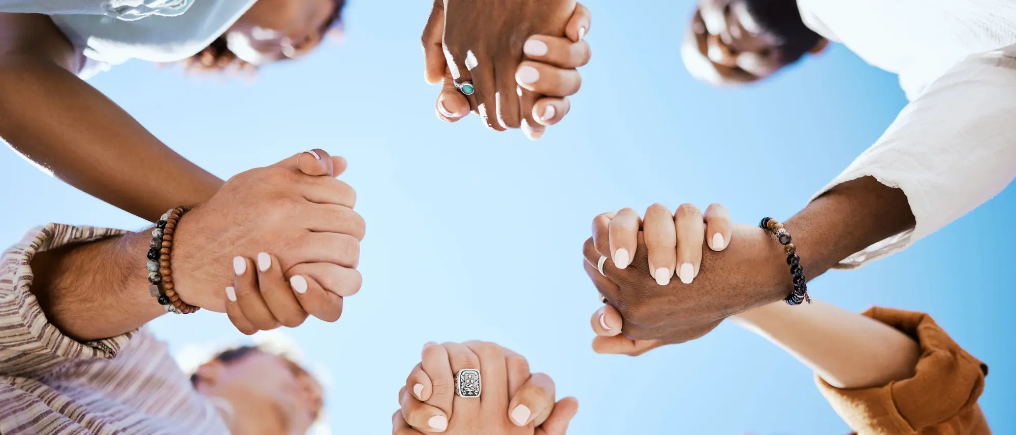 Eine Gruppe von Menschen ist zu sehen, die sich an den Händen halten und im Kreis stehen. Die Hände sind fest miteinander verschlungen, als Symbol für Gemeinschaft und Zusammenhalt. Der Hintergrund zeigt einen klaren blauen Himmel, was der Szene eine freundliche und optimistische Atmosphäre verleiht.