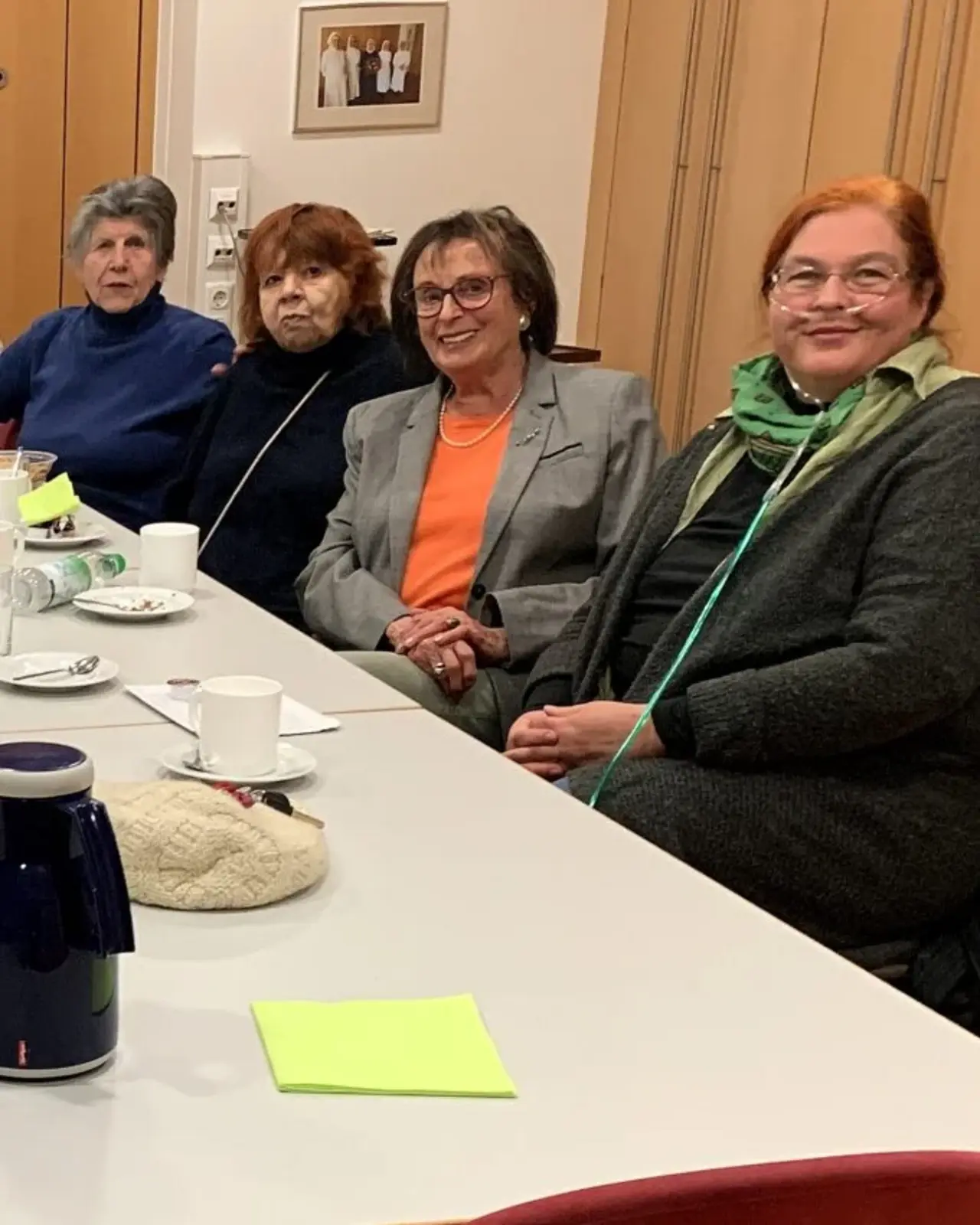 Gesprächskreis: Eine Gruppe von Männern und Frauen unterschiedlichen Alters sitzt um einen großen rechteckigen Tisch in einem Raum. Auf dem Tisch stehen Kaffeetassen, Wassergläser, Flaschen, und Teller mit Gebäck. Die Atmosphäre wirkt entspannt und gesellig.