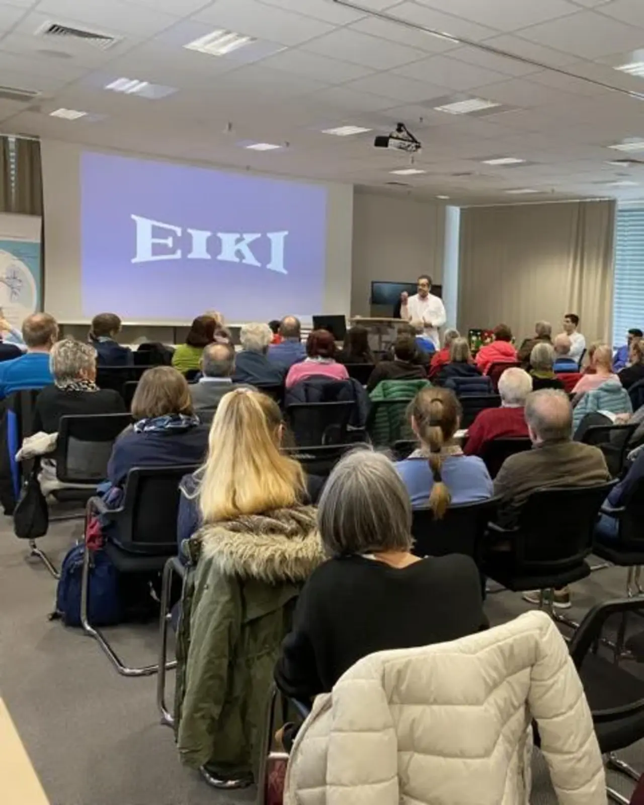 Ein Konferenzraum, in dem fast 50 Personen an einem Treffen in Gießen teilnehmen. Die Zuhörer sitzen in Reihen und schauen nach vorne, wo ein Sprecher neben einer großen Leinwand mit der Aufschrift 'EIKI' steht. Der Raum ist gut beleuchtet und die Teilnehmer hören aufmerksam zu.