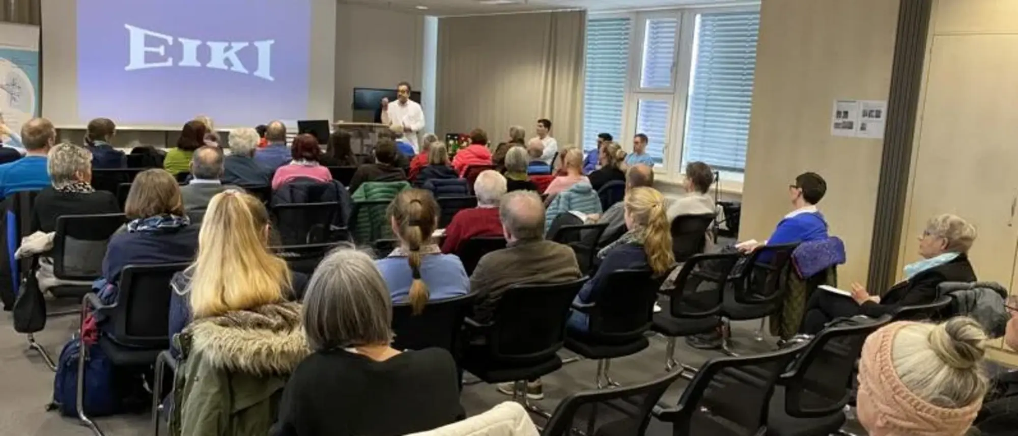 Ein Konferenzraum, in dem fast 50 Personen an einem Treffen in Gießen teilnehmen. Die Zuhörer sitzen in Reihen und schauen nach vorne, wo ein Sprecher neben einer großen Leinwand mit der Aufschrift 'EIKI' steht. Der Raum ist gut beleuchtet und die Teilnehmer hören aufmerksam zu.