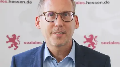 Ein Mann mit Brille und kurzem Haar spricht vor einer Wand mit dem Logo „soziales.hessen.de“. Er trägt ein blaues Sakko und ein hellblaues Hemd.