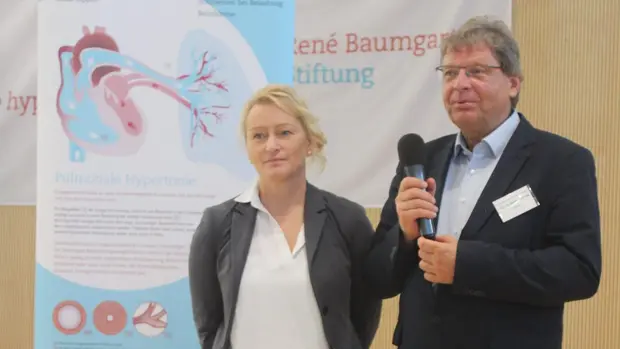 Das Bild zeigt Ekkehard Grünig und Daniela Moritz. Sie stehen nebeneinander auf einer Bühne, während Ekkehard Grünig ein Mikrofon hält. Im Hintergrund ist ein Poster über pulmonale Hypertonie zu sehen.