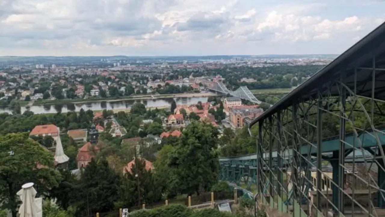 Panoramablick über eine Stadt mit Fluss, Brücke und zahlreichen Häusern, unter einem teils bewölkten Himmel. Im Vordergrund ist ein metallisches Konstrukt zu sehen.