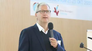 Das Bild zeigt Prof. Dr. med. Dirk Skowasch während einer Präsentation beim 23. PH-Patiententreffen. Er steht an einem Rednerpult, hält ein Mikrofon und spricht zum Publikum. Im Hintergrund ist das Banner der Veranstaltung sichtbar.