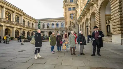 Eine Gruppe von Menschen steht in einem historischen Innenhof, umgeben von prächtigen Gebäuden mit Bögen und Skulpturen. Ein Mann in einem schwarzen Frack und Zylinderhut spricht zur Gruppe, während die anderen aufmerksam zuhören. Eine Frau hält ein dekoratives Netz mit bunten Stoffteilen.