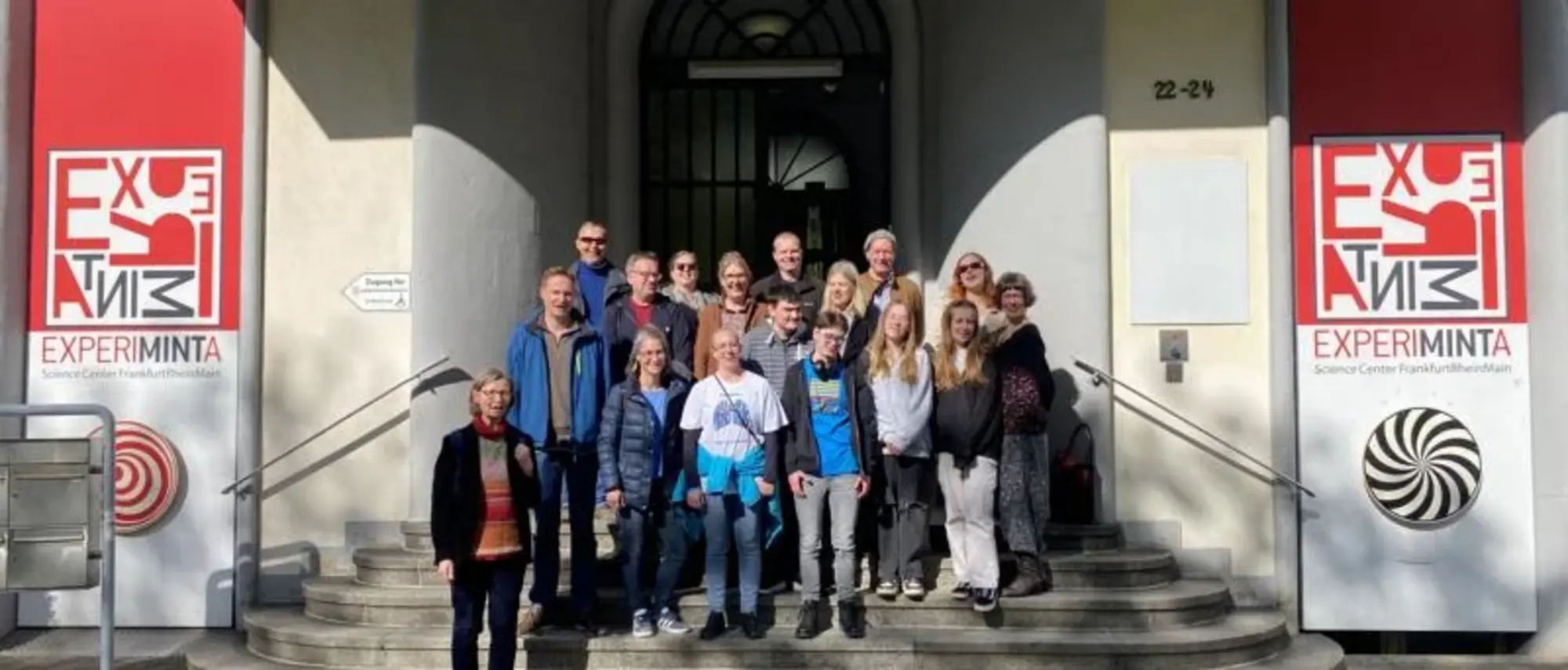 Eine Gruppe von Menschen steht auf den Stufen vor dem Eingang des Experiminta Science Centers in Frankfurt am Main. Sie posieren für ein Gruppenfoto bei sonnigem Wetter.