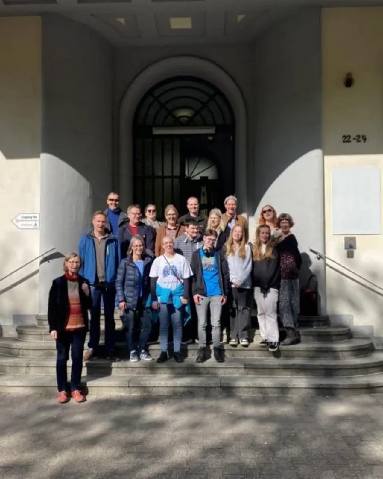 Eine Gruppe von Menschen steht auf den Stufen vor dem Eingang des Experiminta Science Centers in Frankfurt am Main. Sie posieren für ein Gruppenfoto bei sonnigem Wetter.