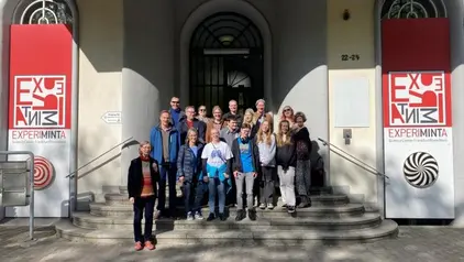 Eine Gruppe von Menschen steht auf den Stufen vor dem Eingang des Experiminta Science Centers in Frankfurt am Main. Sie posieren für ein Gruppenfoto bei sonnigem Wetter.