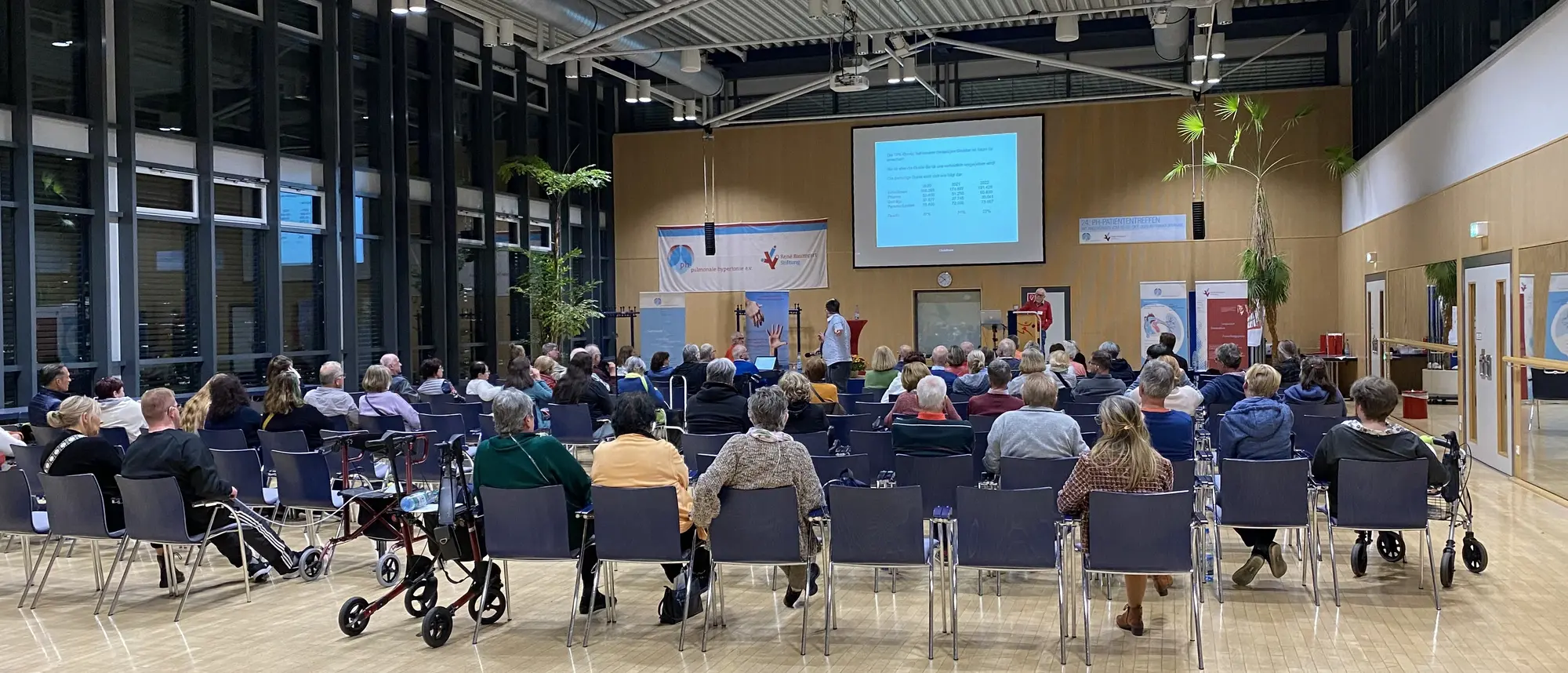 Ein Vortrag im Rahmen eines PH-Patiententreffens, bei dem Zuhörer in einem modernen Veranstaltungsraum einem Redner auf der Bühne zuhören.