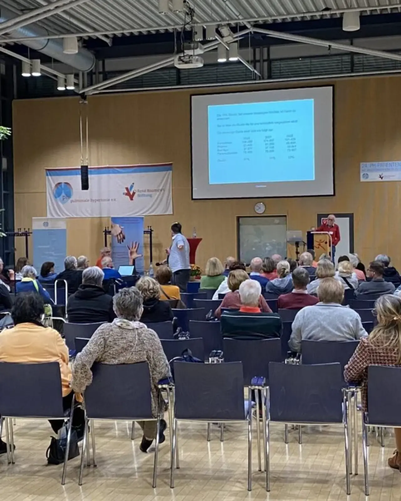 Eine Veranstaltung mit mehreren Teilnehmern in einem großen Saal. Im Vordergrund sitzen Menschen, während im Hintergrund eine Präsentation gezeigt wird.