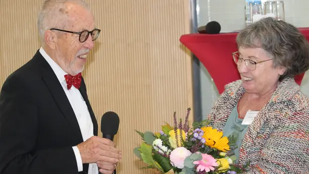 Das Bild zeigt Marlies Schönrock und Hans-Dieter Kulla in einem freudigen Moment. Marlies Schönrock überreicht Hans-Dieter Kulla einen Blumenstrauß, während er ein Mikrofon hält und lächelt.