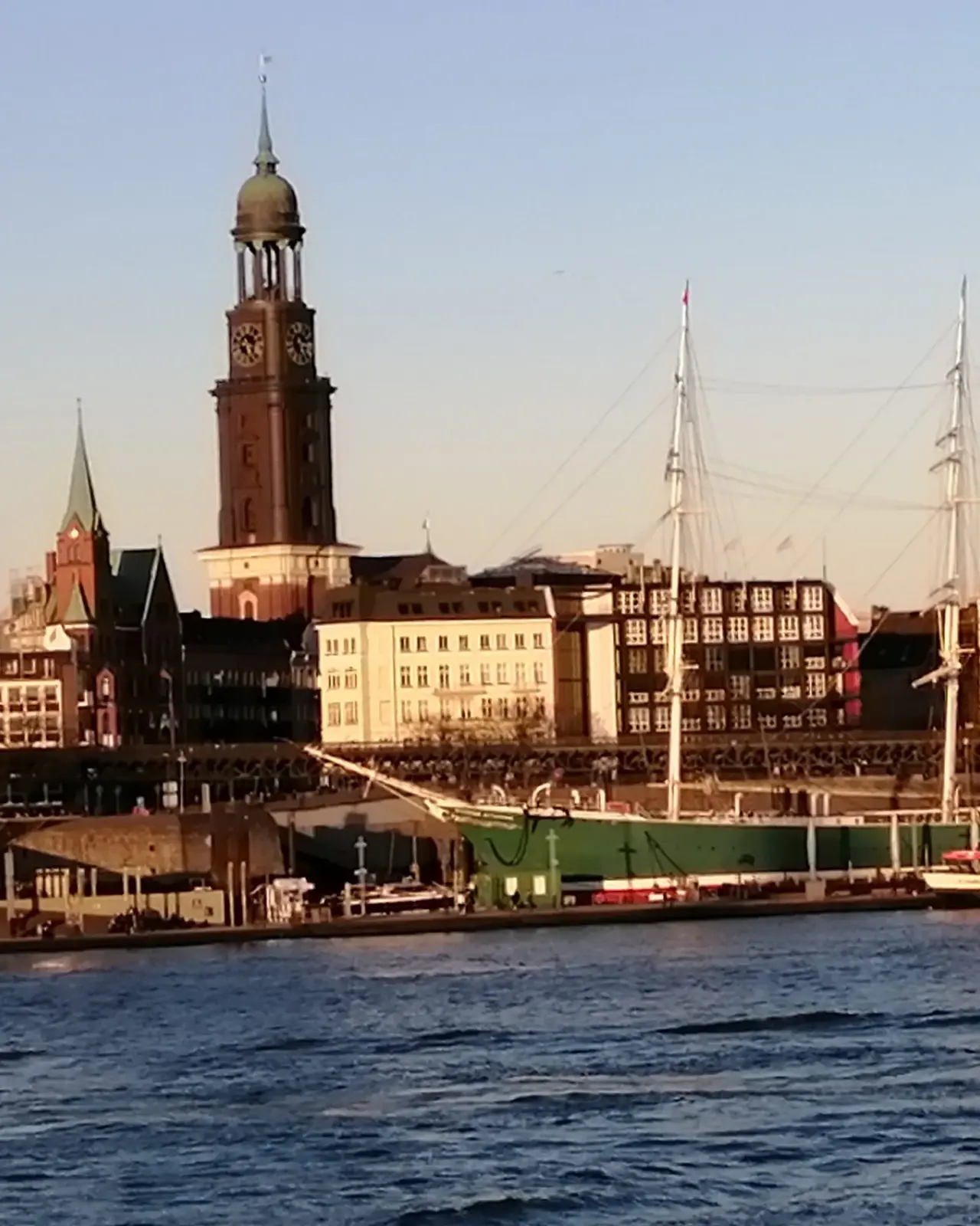 Blick auf den Hamburger Michel, die markante Kirche mit ihrem hohen Turm, im warmen Licht der Abendsonne. Im Vordergrund sind Hafenanlagen und Segelschiffe auf der Elbe zu sehen, die den maritimen Charakter der Stadt unterstreichen.