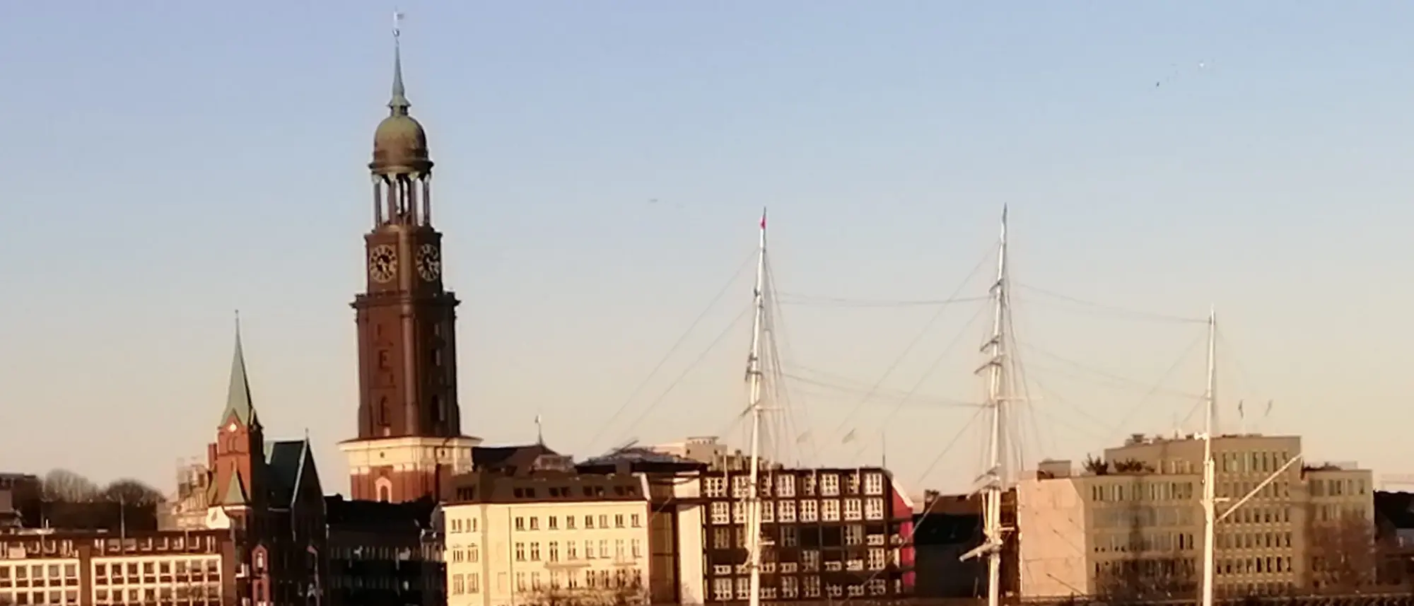 Blick auf den Hamburger Michel, die markante Kirche mit ihrem hohen Turm, im warmen Licht der Abendsonne. Im Vordergrund sind Hafenanlagen und Segelschiffe auf der Elbe zu sehen, die den maritimen Charakter der Stadt unterstreichen.