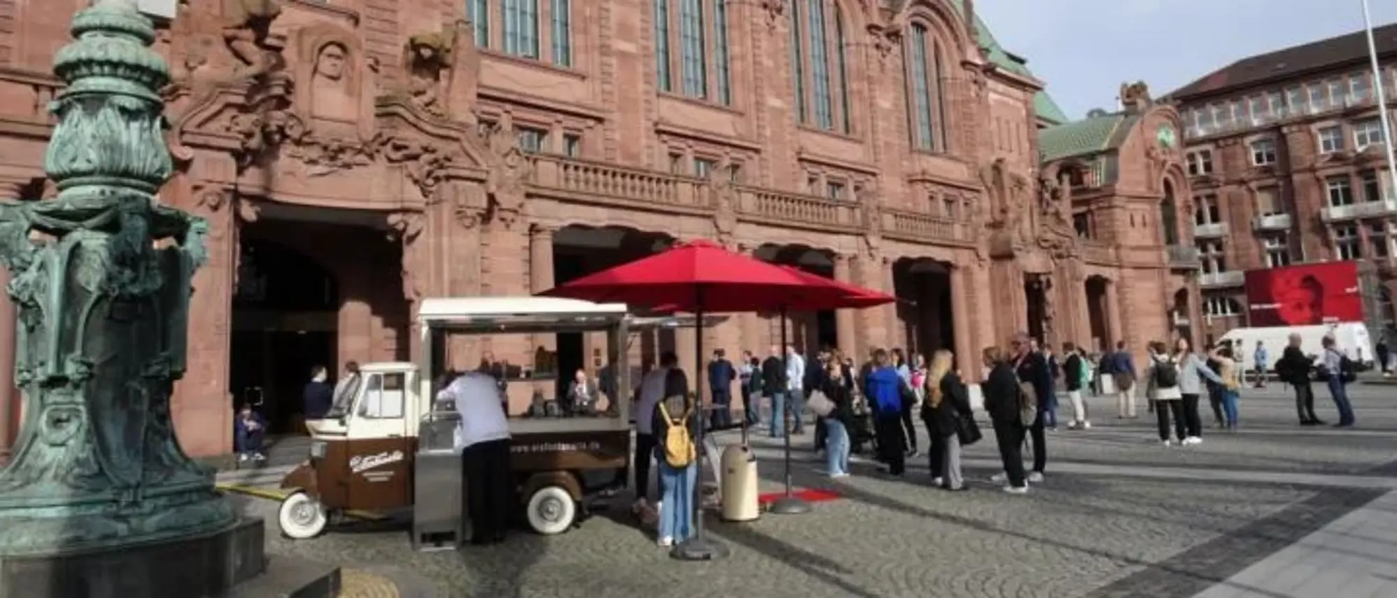 Eine historische, imposante Fassade eines Gebäudes aus rotem Sandstein, vermutlich ein Bahnhof oder ein öffentliches Gebäude. Vor dem Gebäude versammeln sich Menschen auf einem gepflasterten Platz. Ein kleiner Kaffeestand mit einem roten Sonnenschirm steht im Vordergrund, umgeben von Personen, die Getränke bestellen. 