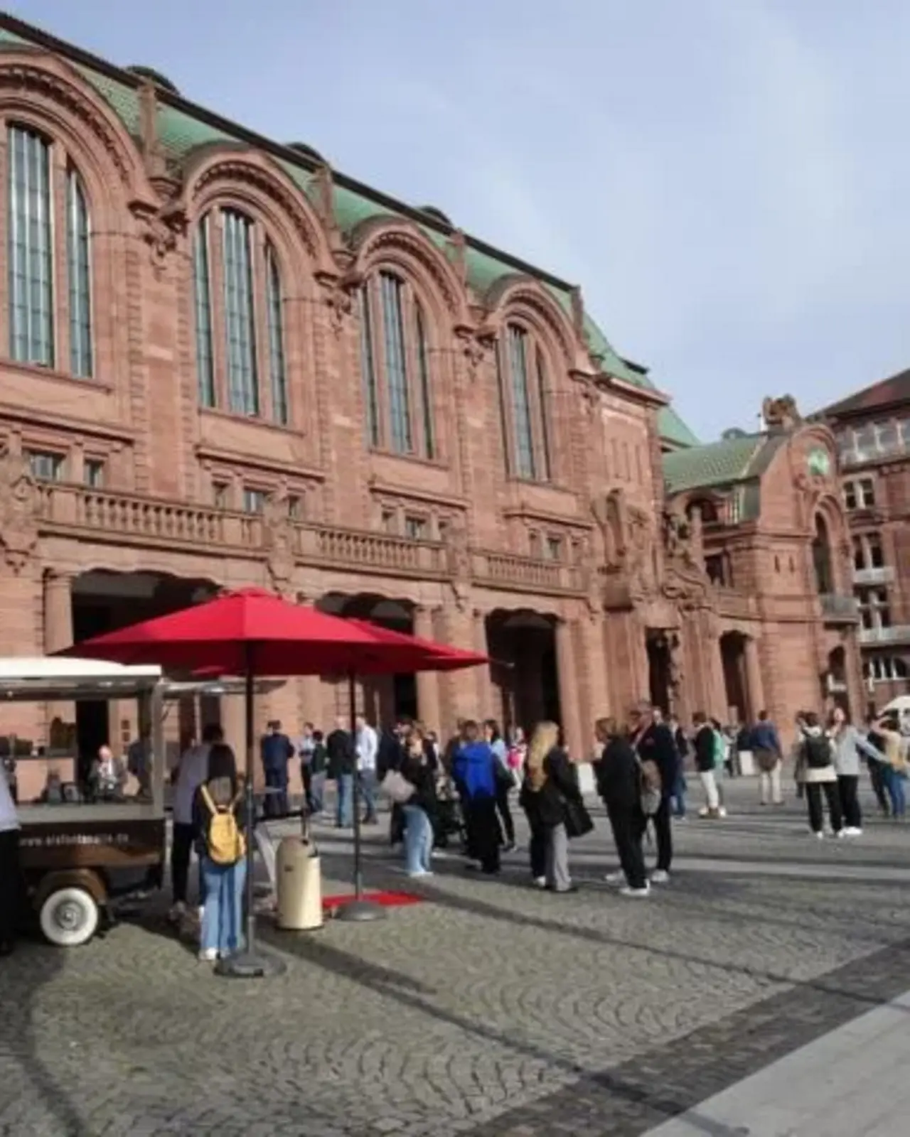 Eine historische, imposante Fassade eines Gebäudes aus rotem Sandstein, vermutlich ein Bahnhof oder ein öffentliches Gebäude. Vor dem Gebäude versammeln sich Menschen auf einem gepflasterten Platz. Ein kleiner Kaffeestand mit einem roten Sonnenschirm steht im Vordergrund, umgeben von Personen, die Getränke bestellen. 