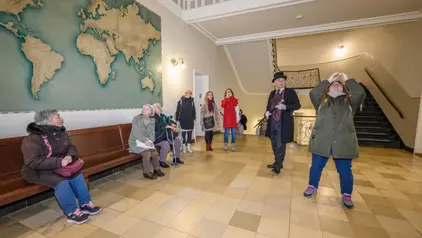 Eine Gruppe von Menschen steht in einer Halle mit einer großen Weltkarte an der Wand. Einige Personen sitzen auf einer Holzbank, während andere stehen und nach oben schauen. Ein Mann in einem schwarzen Frack und Zylinderhut hält einen Spazierstock. Eine Frau macht ein Foto mit ihrem Smartphone. Im Hintergrund sind eine Treppe und ein Geländer zu sehen.