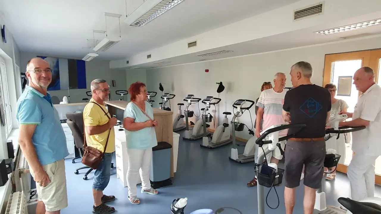 Mehrere Personen stehen in einem hellen Raum mit vielen Fitnessgeräten, wie zum Beispiel Fahrradergometern. Einige der Menschen unterhalten sich miteinander, während andere lächeln. Im Hintergrund sind die Geräte aufgereiht und der Raum ist gut beleuchtet.