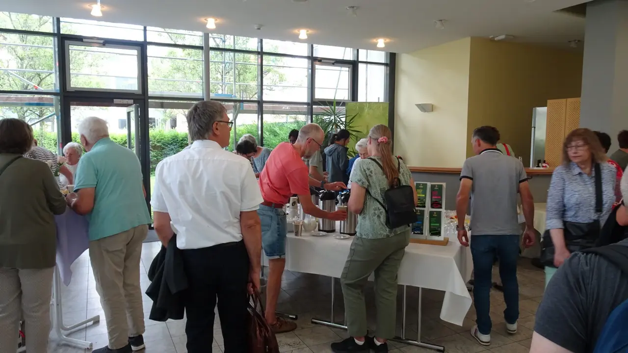 Menschen versammeln sich an einem Buffet-Tisch in einem hellen, modernen Raum mit großen Fenstern. Auf dem Tisch stehen Kaffeekannen und Getränke. Die Teilnehmer bedienen sich und unterhalten sich miteinander. Im Hintergrund sind grüne Pflanzen und eine offene Eingangstür zu sehen, die den Blick nach draußen freigibt.