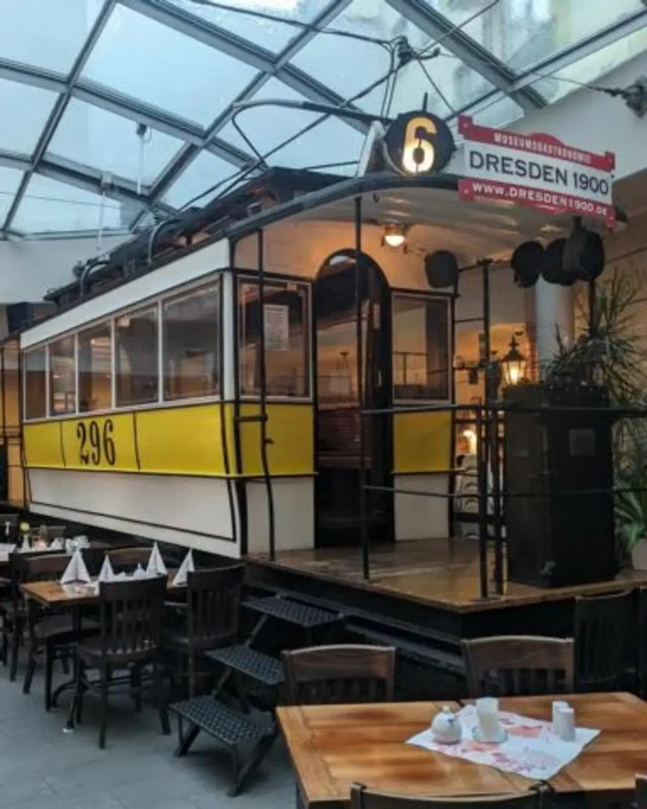 Innenaufnahme eines Restaurants mit einer historischen Straßenbahn als zentrales Dekorationselement. Die Straßenbahn ist gelb-weiß und trägt die Nummer 296. Darüber hängt ein Schild mit der Aufschrift "Dresden 1900". Um die Straßenbahn herum sind Tische und Stühle angeordnet, die für Gäste eingedeckt sind. Das Restaurant befindet sich unter einem Glasdach, das für viel natürliches Licht sorgt. Die Atmosphäre wirkt nostalgisch und gemütlich.